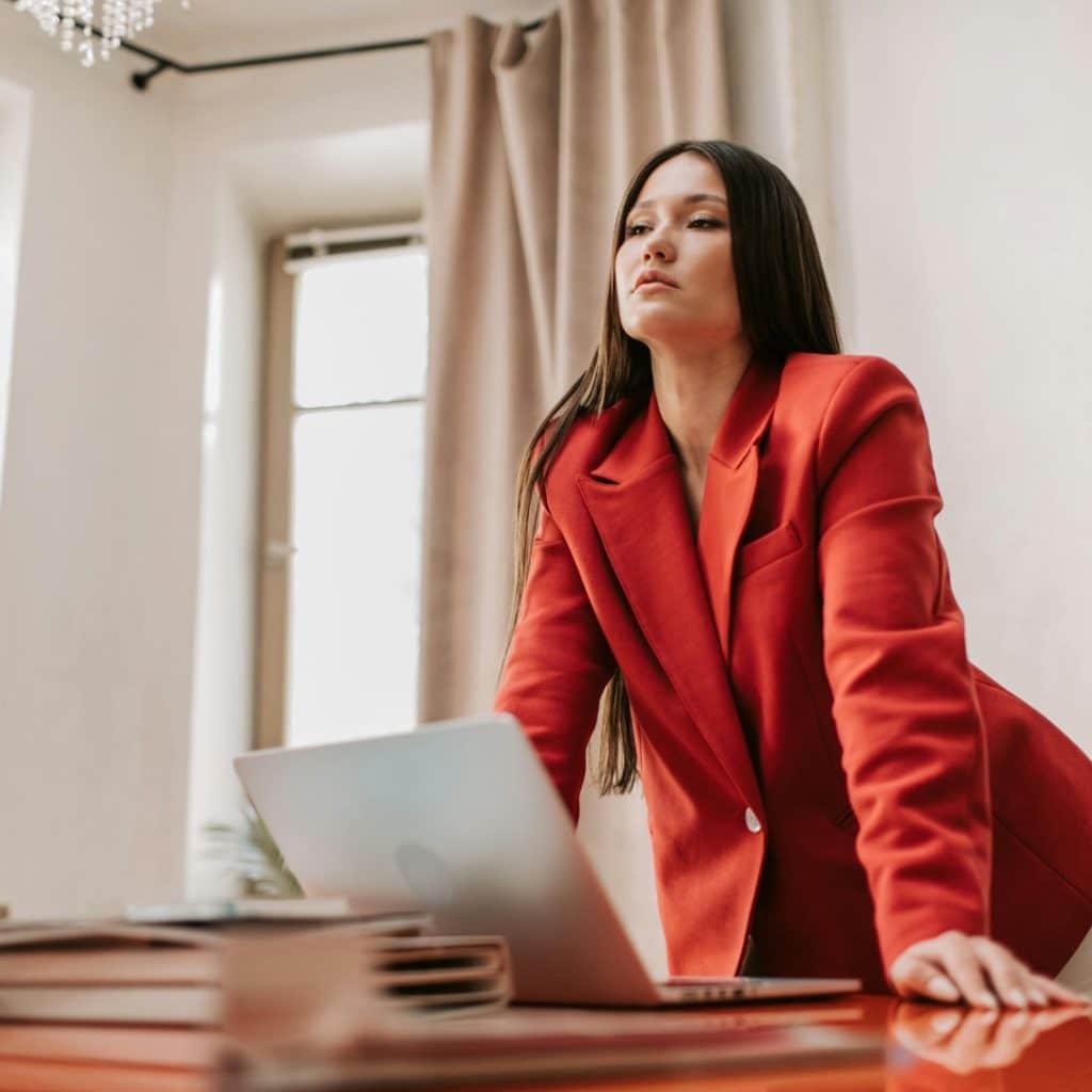 Une femme très chic, qui se tient debout devant un bureau avec son ordinateur. Elle a une posture de femme de pouvoir.